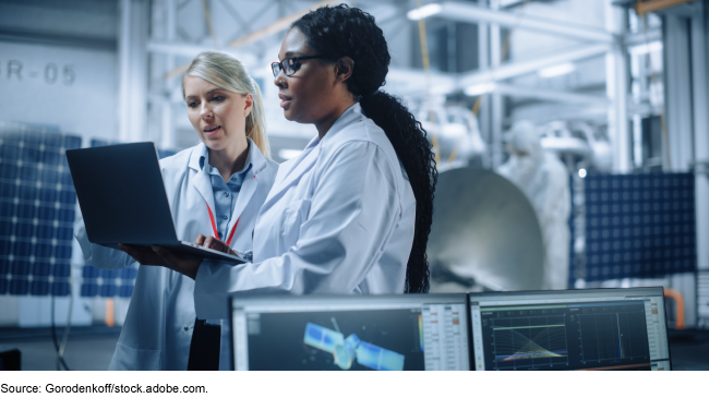 Researchers standing in a lab, surrounded by electrical and other equipment, looking at a laptop. Researchers standing in a lab, surrounded by electrical and other equipment, looking at a laptop.