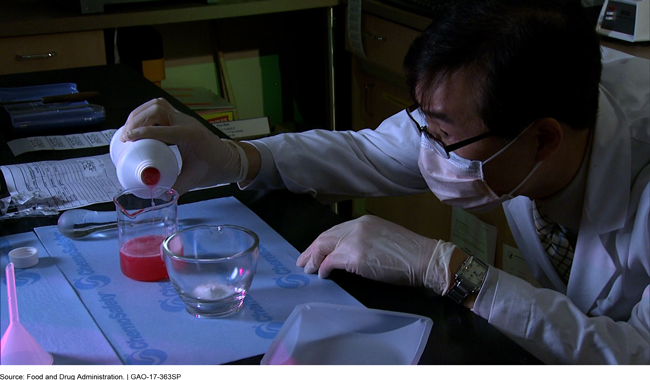 Photo of pharmacist, wearing a mask and gloves, pouring red liquid into a glass beaker.