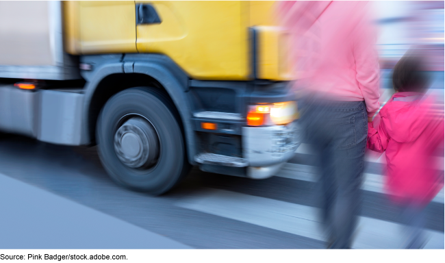 An adult holding the hand of a young child walking on a pedestrian crossing with a large vehicle closely approaching in front of them. An adult holding the hand of a young child walking on a pedestrian crossing with a large vehicle closely approaching in front of them.