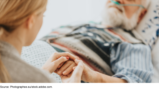 A woman holding the hand of an older man who is receiving supplemental oxygen from a medical device while in bed.