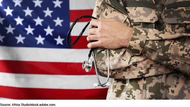 Close up of a person’s torso and arms wearing camouflage holding a stethoscope. An American flag is in the background.