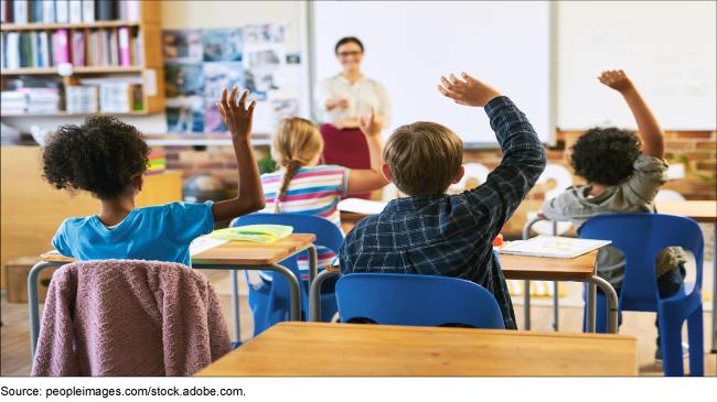 Multiple children with their hand raised looking towards an adult pointing in a classroom setting.