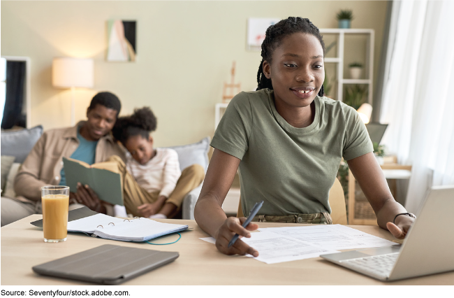 A woman in a military uniform sits in front of a laptop computer, and in the background a man reads to a young girl. 