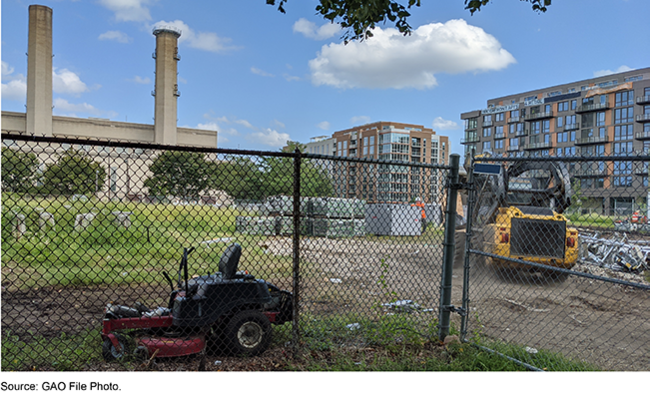 Chain link fence around construction equipment, materials and debris with buildings in the background.