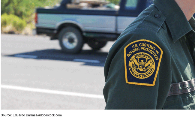 Customs and border patrol patch on the sleeve of an officer standing outside with a pickup truck in the background.