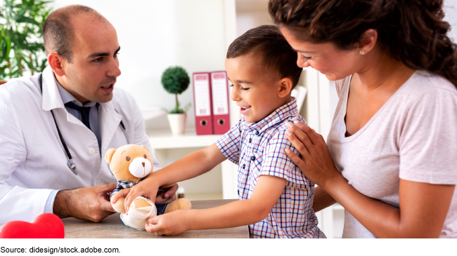 Photo showing a doctor with a small child and his mother. The child is playing with a teddy bear, which has a bandage wrapped around its foot.