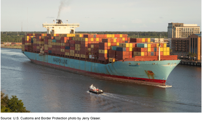 A large cargo ship transporting stacks of shipping containers on a body of water, with buildings and trees on the shoreline behind the ship.