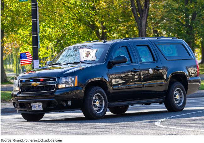 A dark colored SUV driving with two small flags on the front and blue and red lights in the front grill.