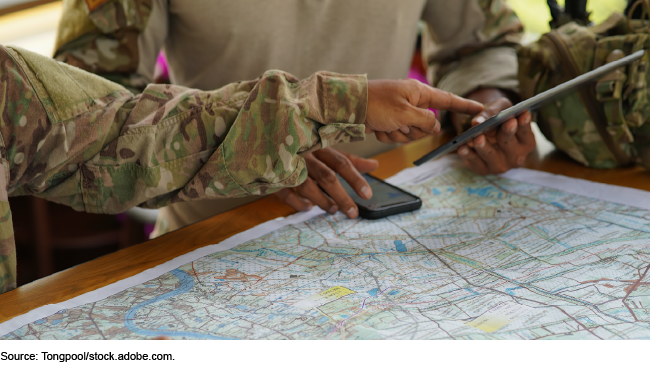 Two people in camouflage clothing pointing at a tablet while viewing a map and a phone on a table Two people in camouflage clothing pointing at a tablet while viewing a map and a phone on a table