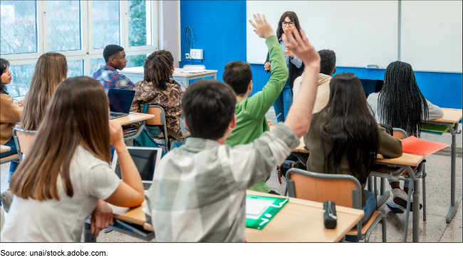 Photo from a classroom where two students raise their hands and the rest focus on a person in front.