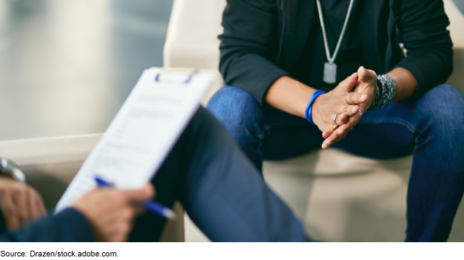 A person wearing military dog tag seated across from a person with a clipboard.