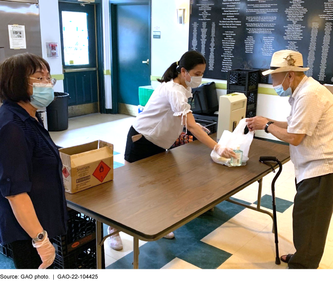 A woman wearing a mask hands a bag of food across a table to an elderly man also wearing a mask.