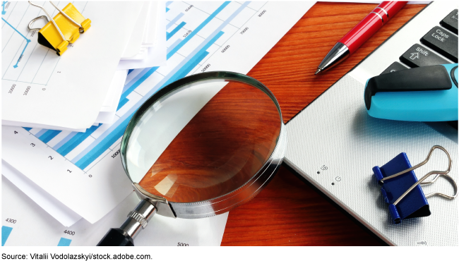 A magnifying glass lying on a desk surrounded by a laptop computer, papers, and pens.