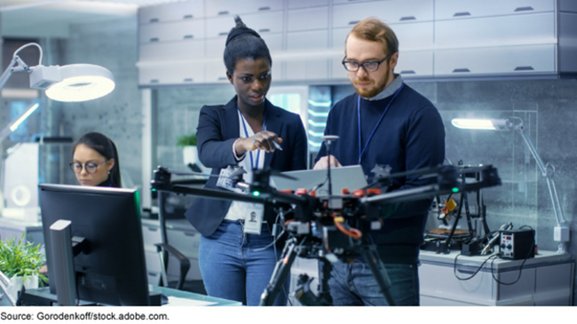 Three people in a technology lab. Two are discussing a drone-type item that is on the table in front of them.