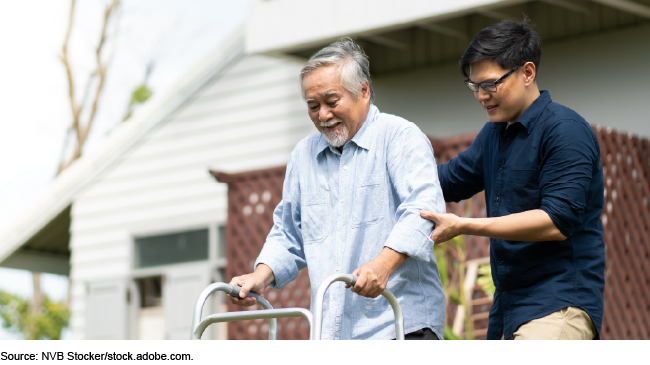 A younger man helps an older man to use a walker in the back yard of a house.