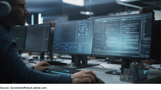 A man wearing headphones works at a computer keyboard in front of three monitors displaying computer code and other data.