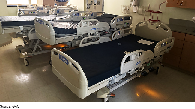 A room containing 3 empty hospital beds with bare mattresses and some cardboard boxes full of stuff.