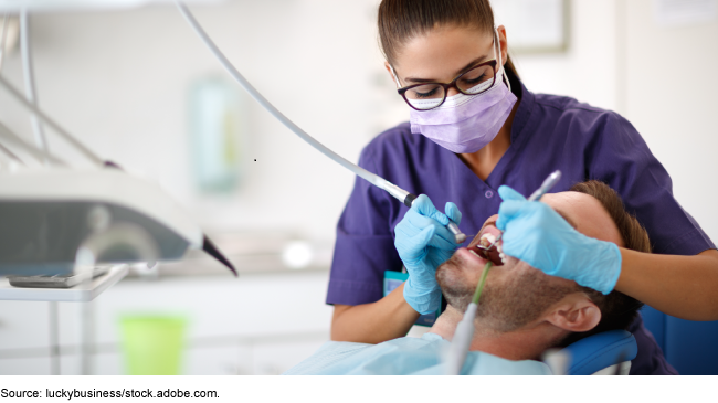 A woman in a medical mask and glasses holds dentist tools with rubber-gloved hands and puts the tools in a man's mouth.