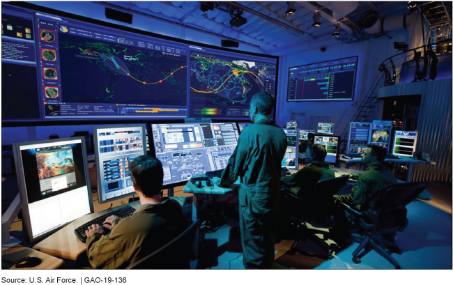 Military personnel looking at a bank of large monitors in a dimly lit room.