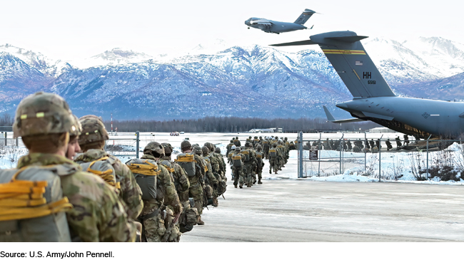 Army paratroopers lining up to board a large transport aircraft in front of snowy mountains.