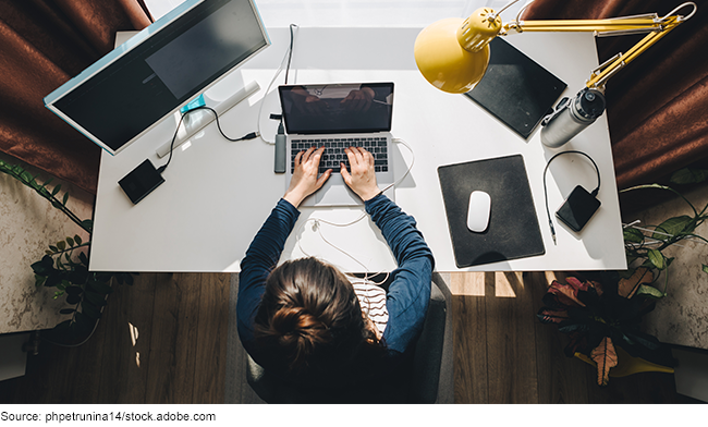 A female employee is working on a laptop and monitor at her desk at home.