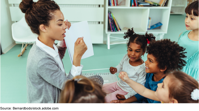 A teacher and children sitting on the ground while the teacher points to a notebook.