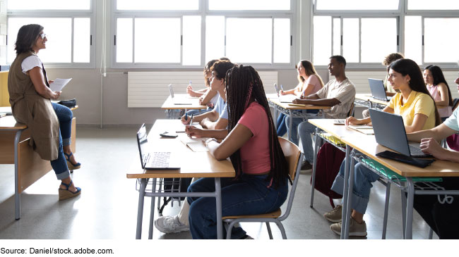 A classroom setting with young people sitting at desks looking at an adult holding a piece of paper at the front of the room.