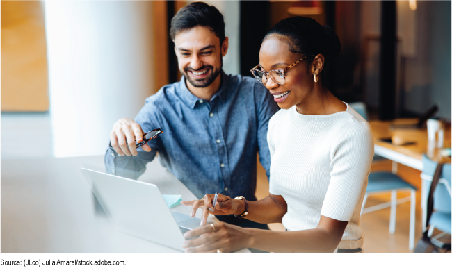Two people at a desk using a laptop in a bright office setting. 