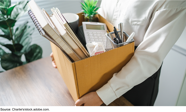 A person wearing a button-down shirt stands in an office, holding a cardboard box filled with various office supplies and a small plant.