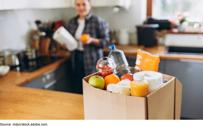 A person in a corner of an apartment kitchen, standing next to a glass stovetop, with countertop appliances on either side. A cardboard box filled with groceries and paper towels is sitting on the countertop in the foreground.