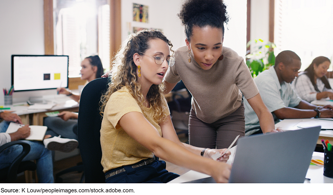A woman sitting at a desk with a laptop in front of her while another woman stands next to her also looking at the laptop. A woman sitting at a desk with a laptop in front of her while another woman stands next to her also looking at the laptop.