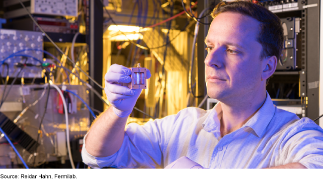 A man in a white shirt and rubber gloves holds a small box with internal circuitry in a room with complex electronic equipment.