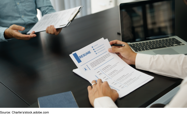 A person reading a resume, sitting across a desk from someone holding a clipboard. A person reading a resume, sitting across a desk from someone holding a clipboard.