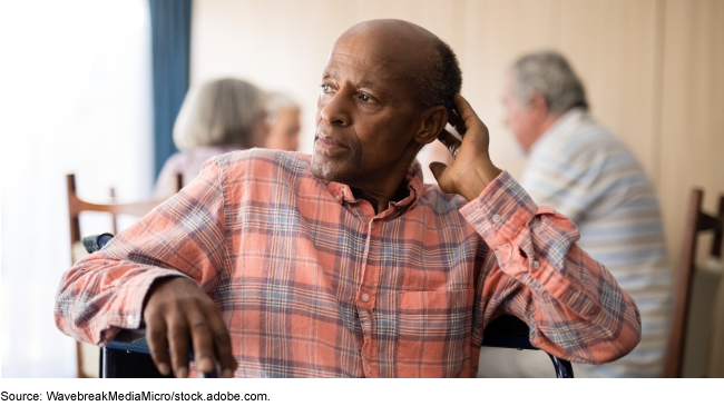 An older person sitting in a chair in a room with other older people in the background. An older person sitting in a chair in a room with other older people in the background.