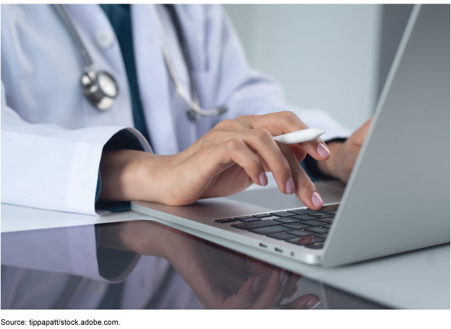  A person wearing a white coat and a stethoscope sitting at a desk typing on a laptop.