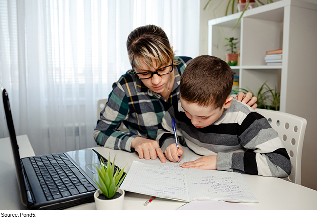 Caregiver sits with a child writing on paper with a laptop on the table.