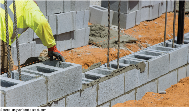 A mason builds a wall by laying a grey, precast concrete block on a mortared row. A mason builds a wall by laying a grey, precast concrete block on a mortared row.