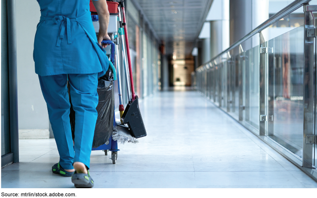 A custodian pushes a cart with cleaning supplies down a corridor.