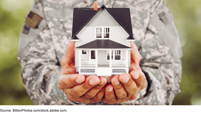 A uniformed servicemember holding a miniature two-story house cupped in both hands with arms stretched out in front of him