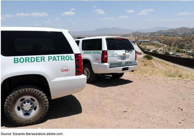 Two white SUVs parked in the dirt next to a road, with a long fence on the right-hand side of the road. Two white SUVs parked in the dirt next to a road, with a long fence on the right-hand side of the road.