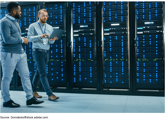 A picture containing two cyber professionals walking in a data center next to server racks while viewing data on a laptop.
