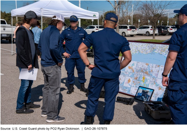 A group of people gathered on an outside parking lot on a sunny day. Coast Guard personnel in dark blue uniforms are talking to people. A large map is propped up on the ground. 