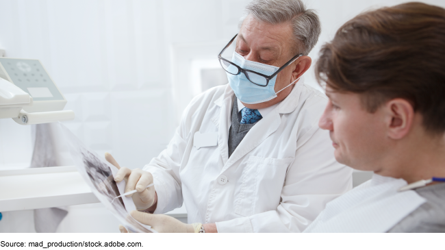 A person wearing glasses and a doctor’s white coat holding papers sitting next to another person sitting in a chair in a dental office setting.