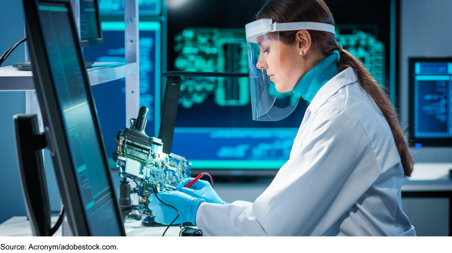 A person in a white coat and a clear face shield works in a laboratory on a circuit board.
