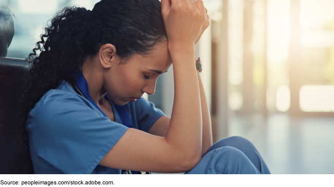 A woman with curly hair, wearing short-sleeved hospital scrubs, sits on the floor with her head in her hands.