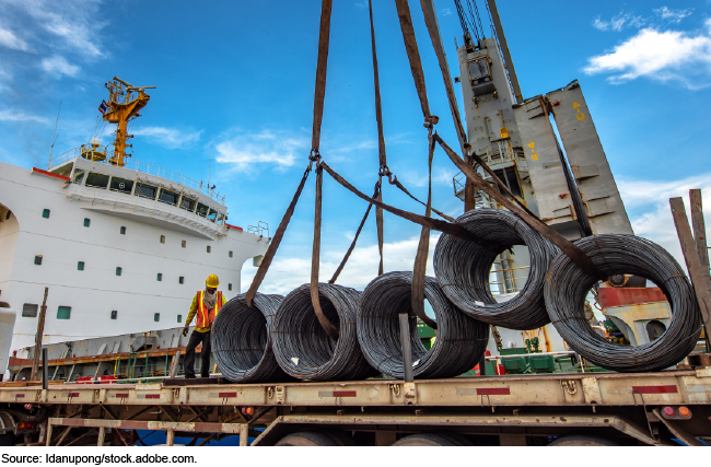 a crane lifting steel coils on a ship, a worker wearing a safety vest and helmet, and the sky in the background