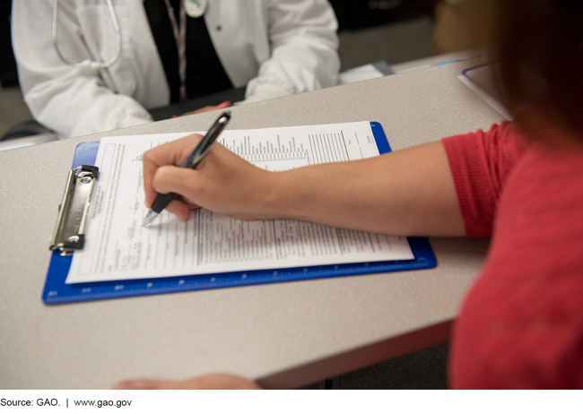 A photo of a patient completing a form on a clipboard in front of a medical professional.