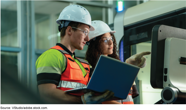 Two people in reflective vests, hard hats, and safety glasses are looking at computers.&nbsp;