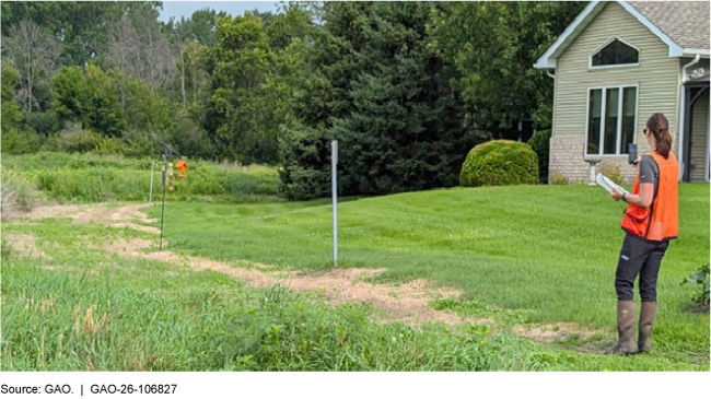 A U.S. Army Corps of Engineers official standing on a pathway in grass next to a home, holding up a handheld device.