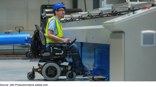 A man in an electric wheelchair, wearing safety glasses, a yellow vest and blue helmet in a factory.